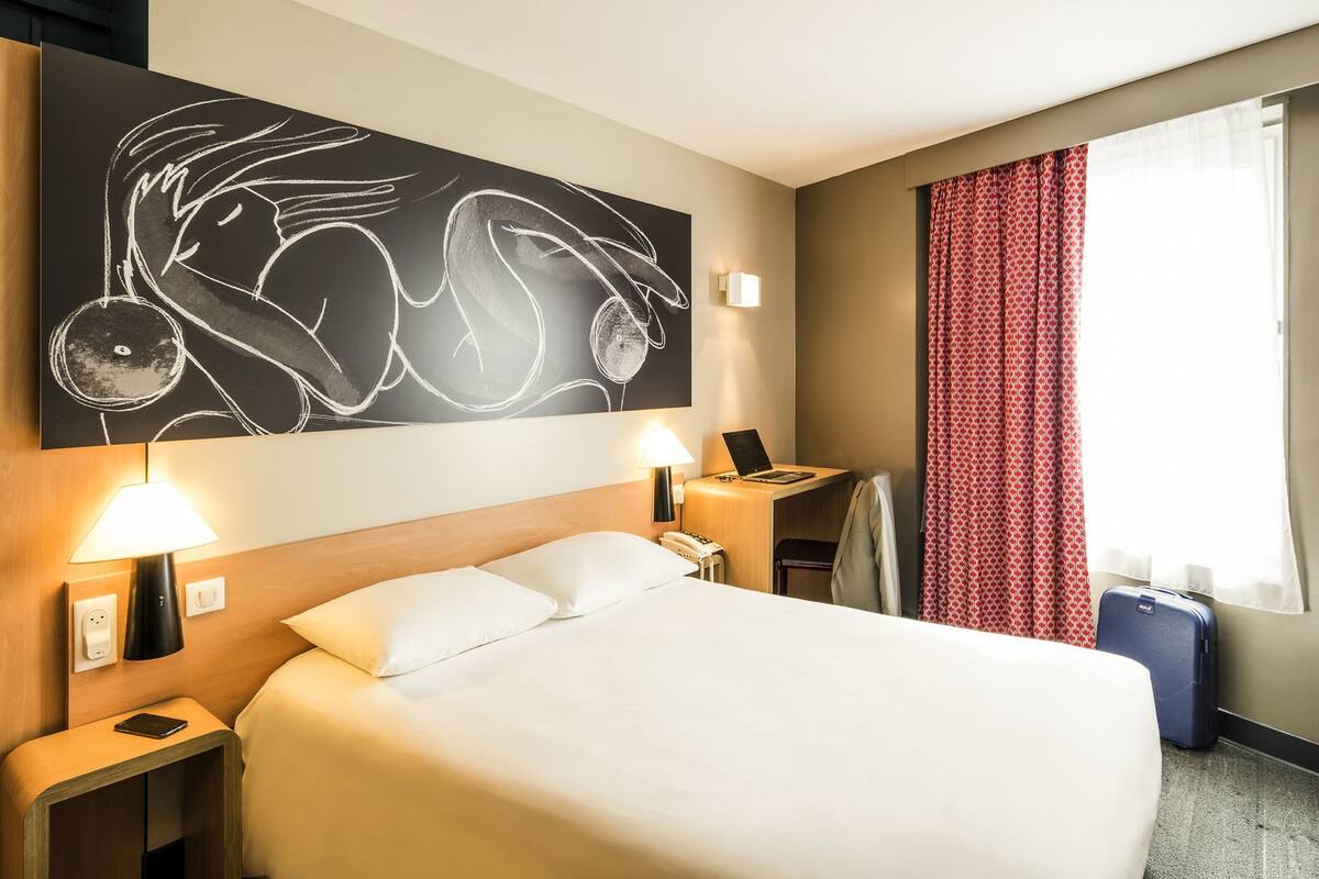 A hotel room with a large bed, bedside lamps, a desk with a laptop, a telephone, and a suitcase.