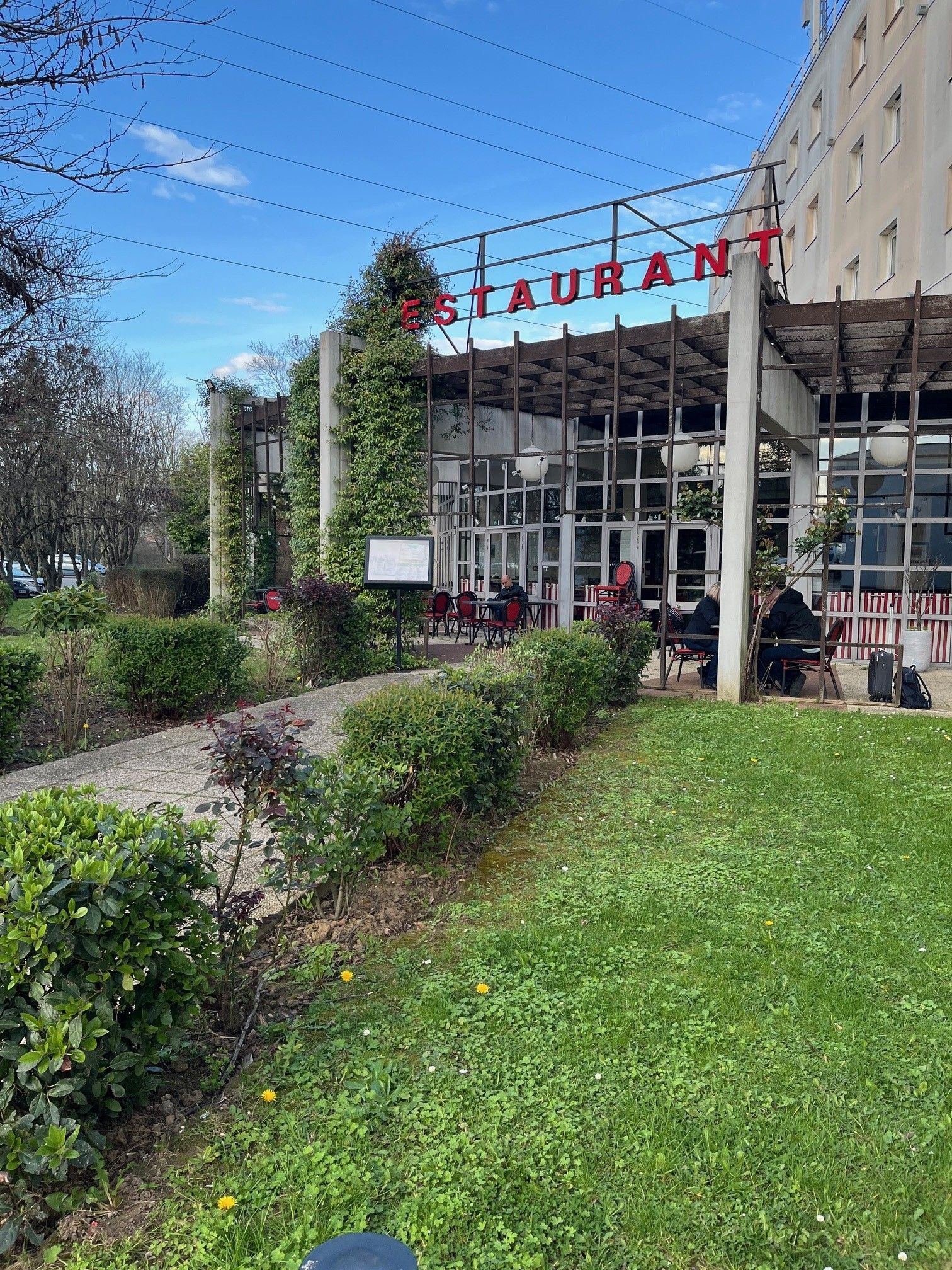 Un restaurante con mesas al aire libre, un letrero sobre la entrada y un jardín con vegetación y un camino.