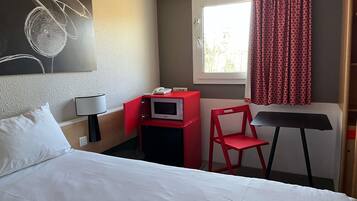 Ein Hotelzimmer mit einem Bett, einem Schreibtisch mit Mikrowelle, einem Sessel, einer Lampe und einem Fenster mit Vorhängen.