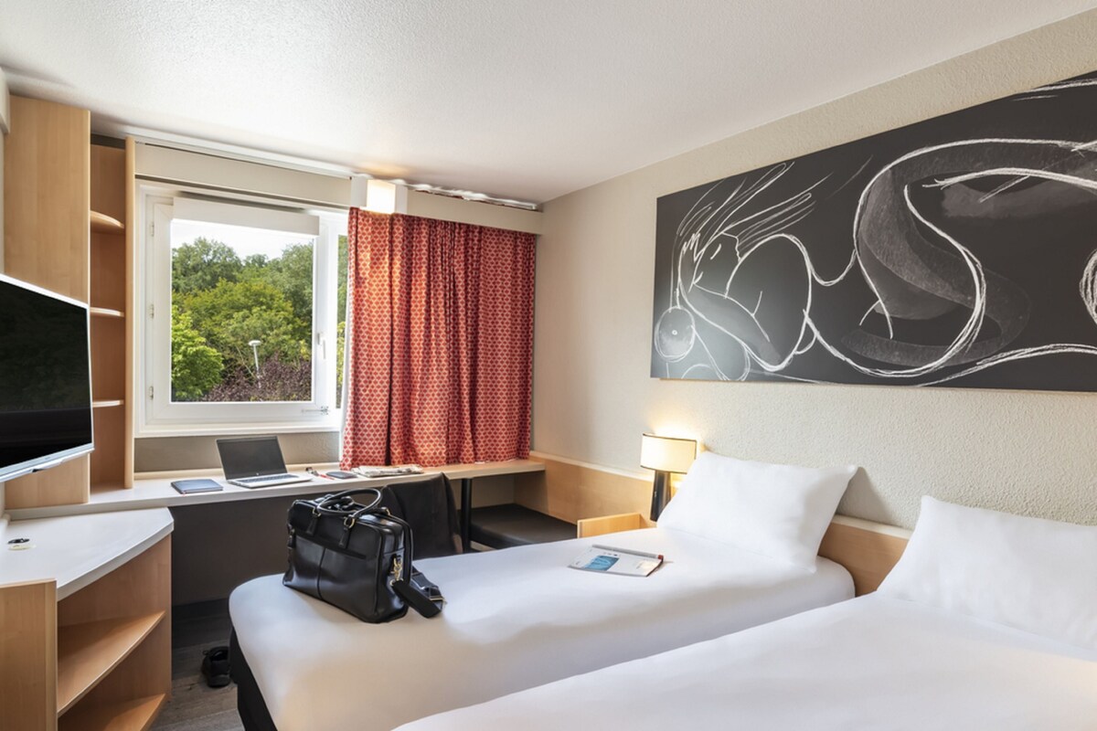 A hotel room with two beds, a desk with a laptop, a TV, and a large abstract painting on the wall.