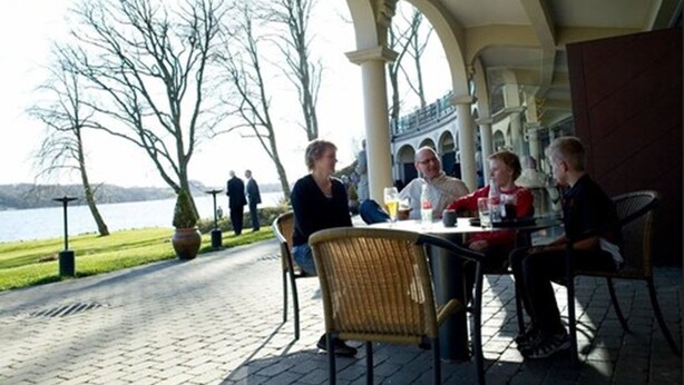 An outdoor dining area with a view of a lake, featuring a brick pathway, wicker chairs, and a table with drinks. - Hotel Koldingfjord (Kolding)