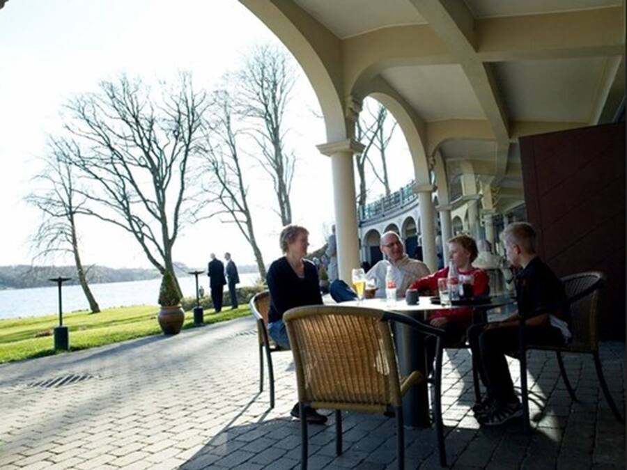An outdoor dining area with a view of a lake, featuring a brick pathway, wicker chairs, and a table with drinks.