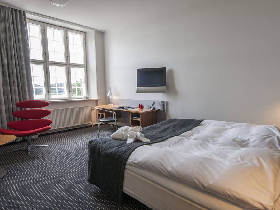 A hotel room with a bed, a desk, a chair, a TV, and a window with curtains.