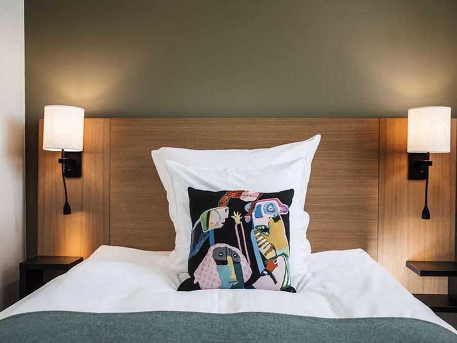 Una cama bien hecha con un cojín colorido, cabecera de madera y dos lámparas de noche.