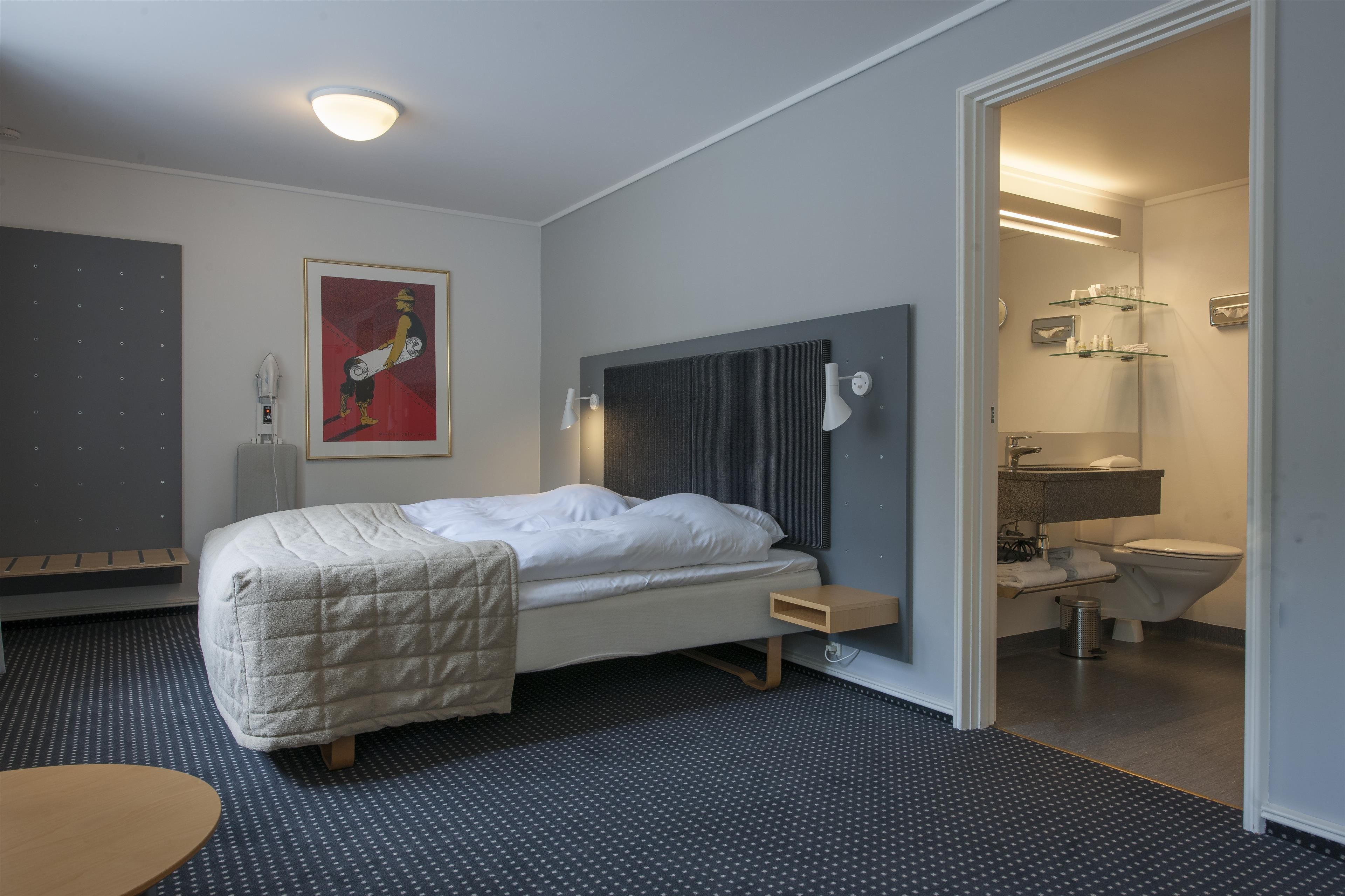 Habitación de hotel moderna con cama, mesita de noche y un baño visible a través de una puerta abierta.