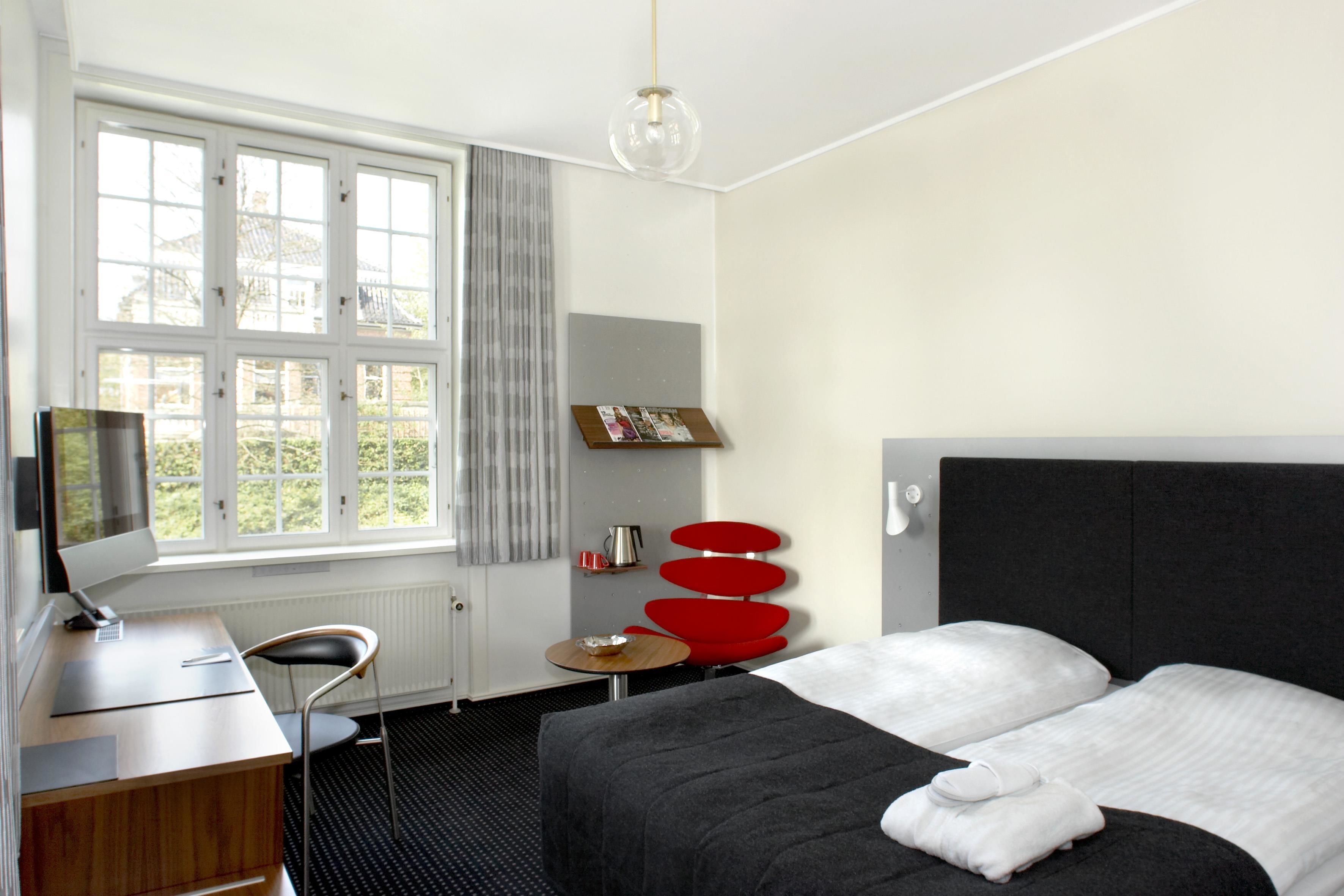 A hotel room with a bed, a desk, a chair, a small table, and a window with curtains.