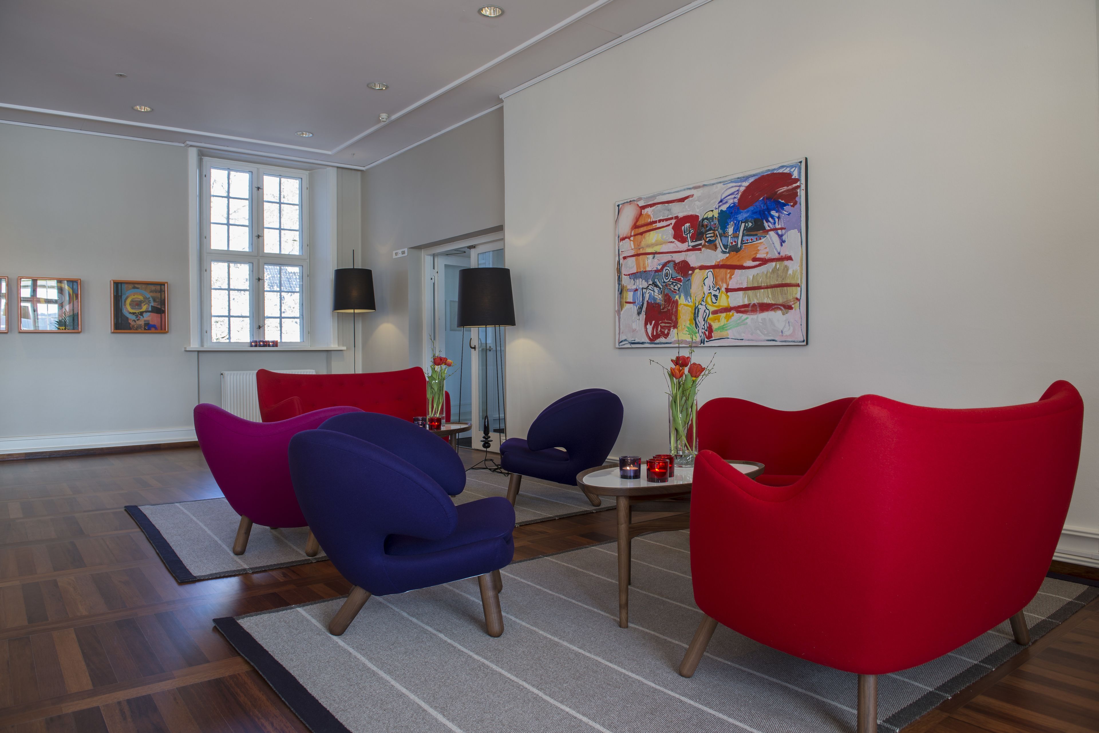 Una sala moderna con sillones rojo y azul, una mesa de centro y un cuadro abstracto grande en la pared.