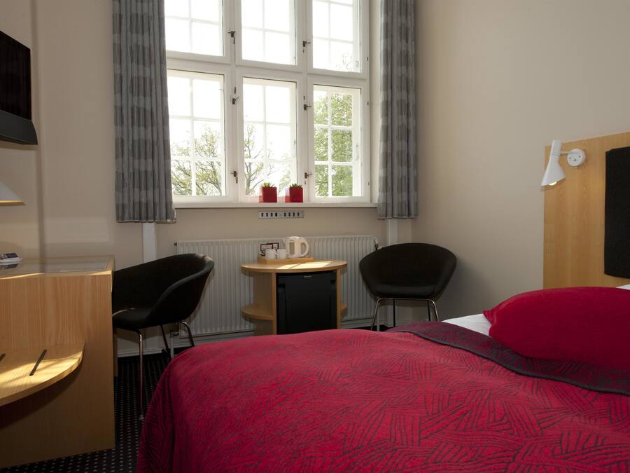 Una habitación de hotel con una cama, un escritorio y una silla, una mesita con una tetera y tazas, un televisor montado en la pared, y una ventana con cortinas.