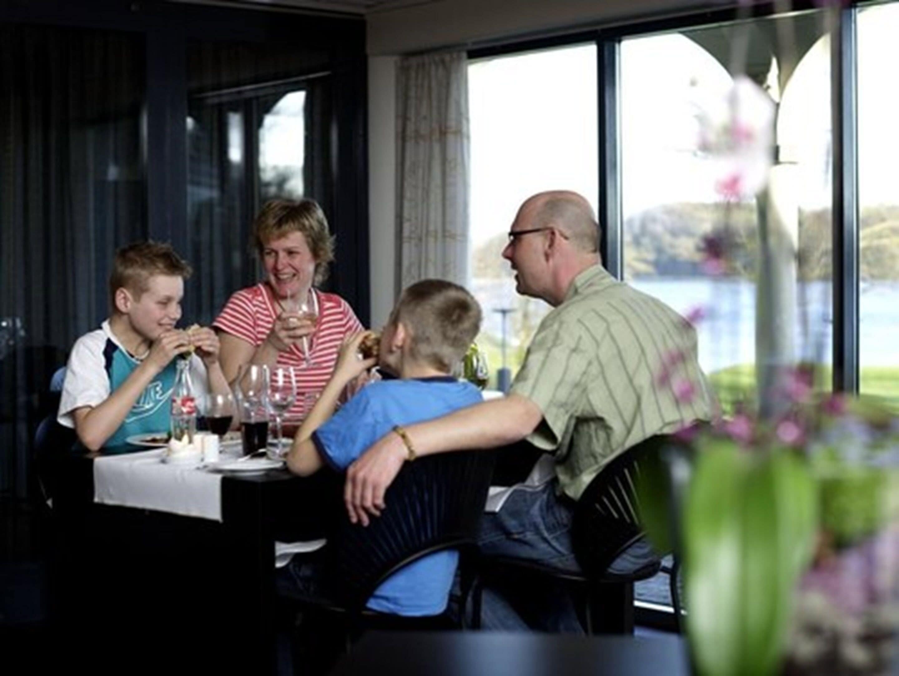 Une famille est assise à une table, près d’une fenêtre donnant sur un plan d’eau, et y prend son repas.
