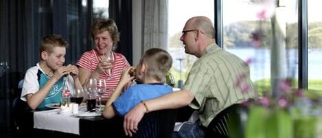 Une famille est assise à une table, près d’une fenêtre donnant sur un plan d’eau, et y prend son repas.