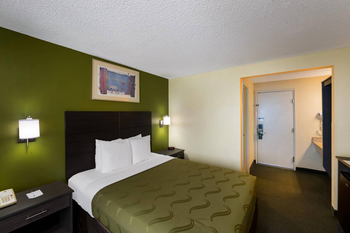 Une chambre d’hôtel comprenant un lit, une table de chevet, une lampe fixée au mur, un tableau et une porte donnant sur une salle de bain.