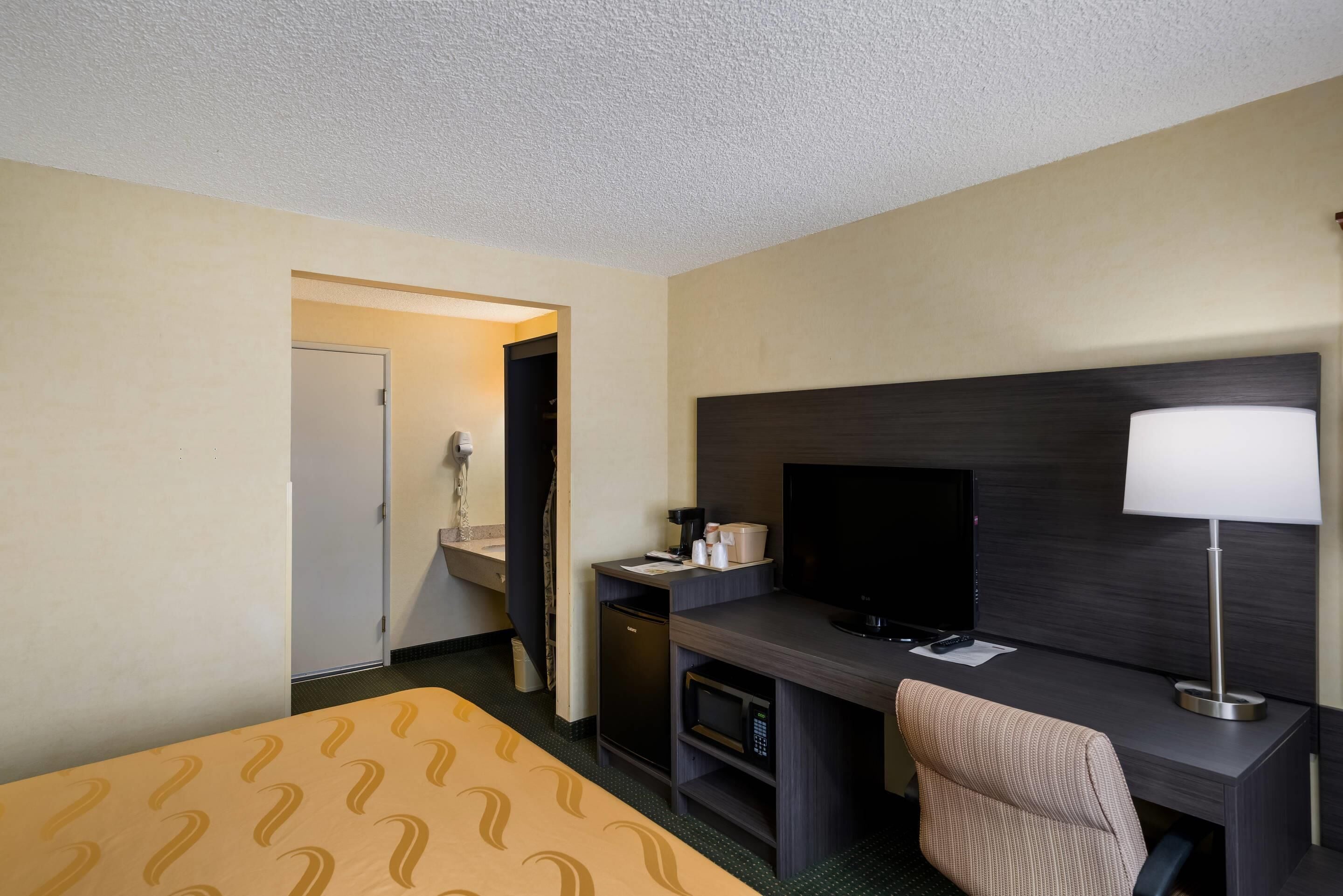 Ein Hotelzimmer mit Bett, Schreibtisch, Fernseher, Lampe und einer kleinen Küchenzeile.