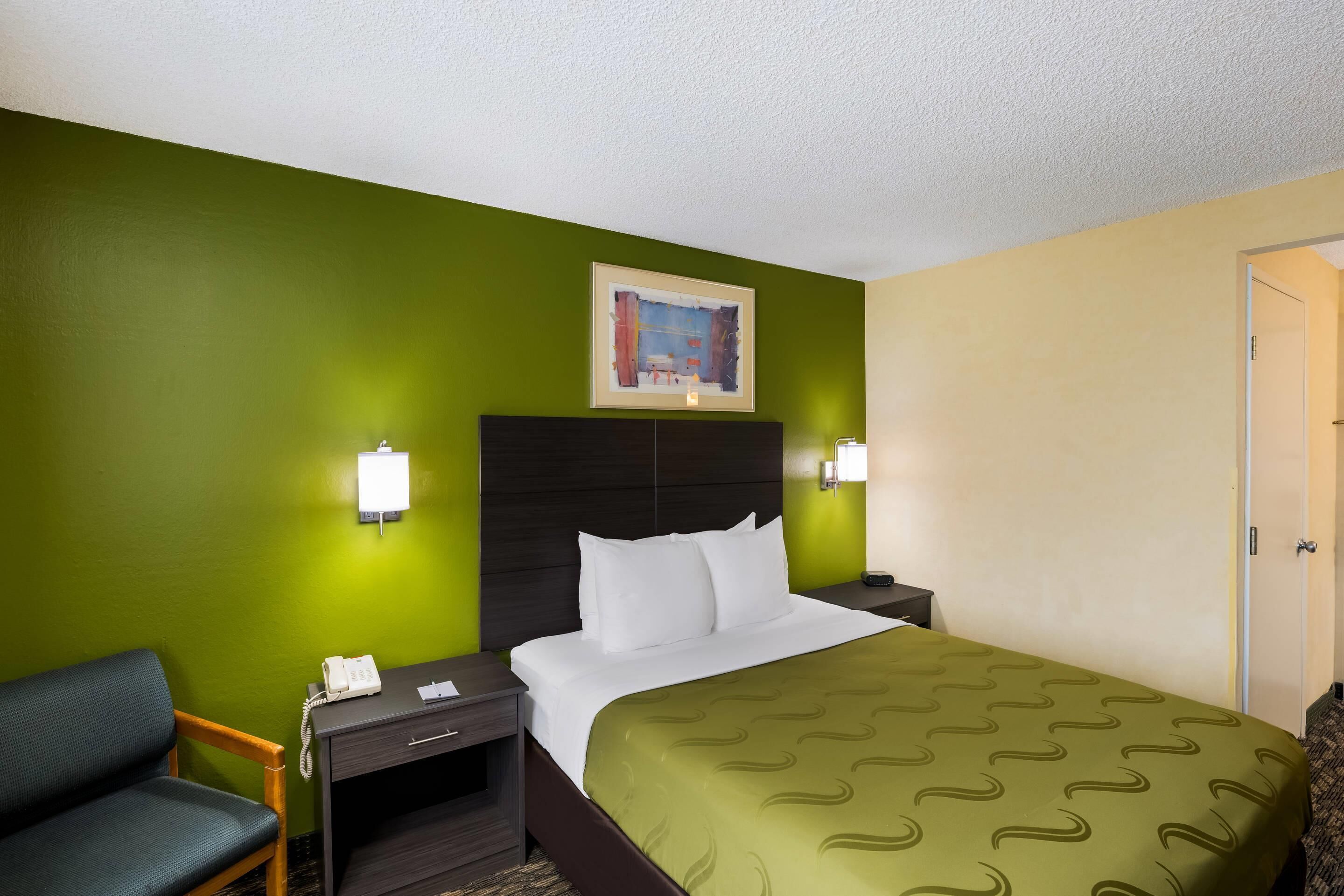 A hotel room with a green wall, a bed with white linens, a dark headboard, and a framed picture above the bed.