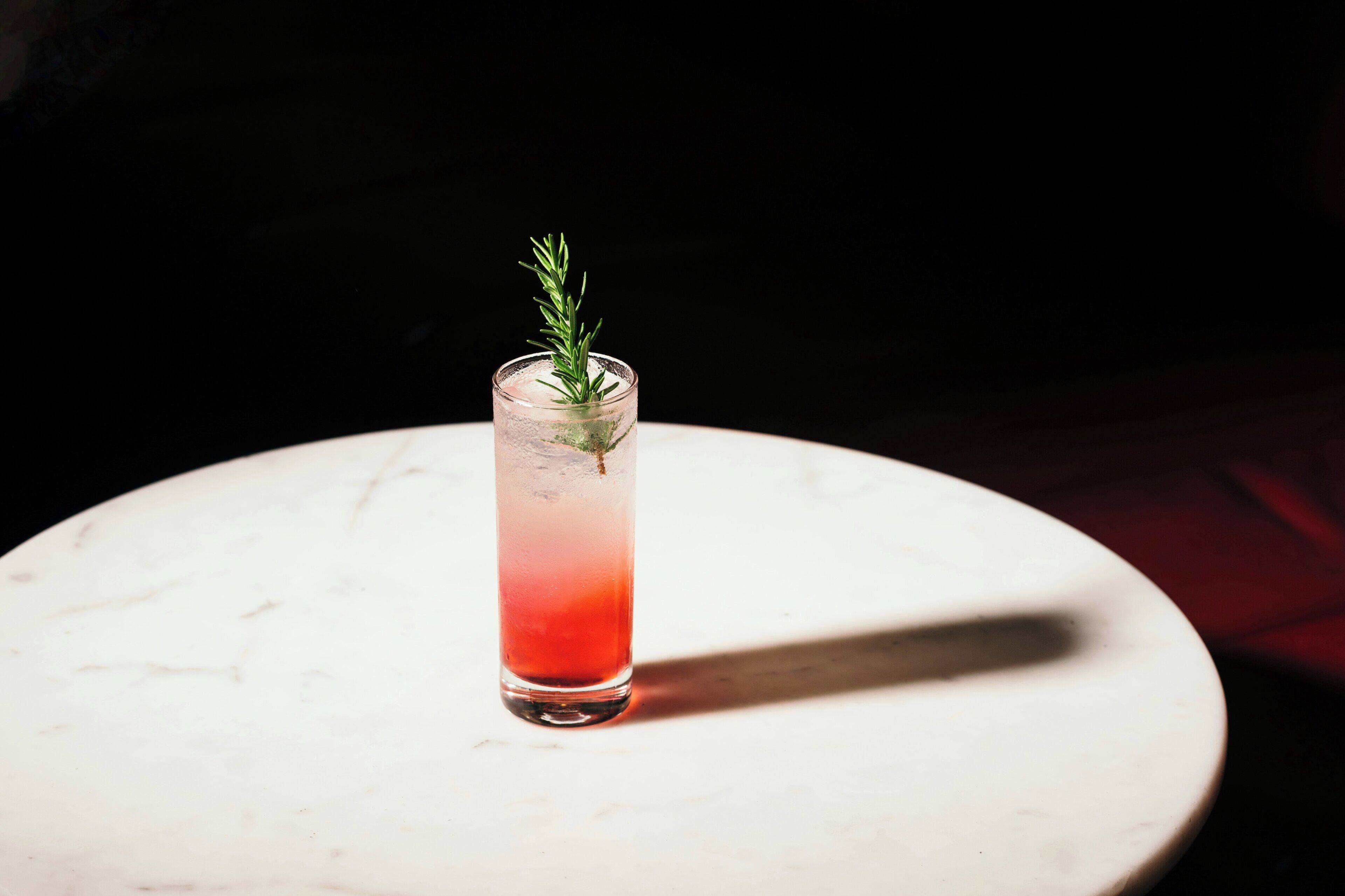 A tall glass with a gradient of red to clear liquid, garnished with a sprig of rosemary, on a white marble surface.