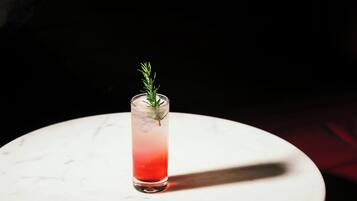 A tall glass with a gradient of red to clear liquid, garnished with a sprig of rosemary, on a white marble surface.
