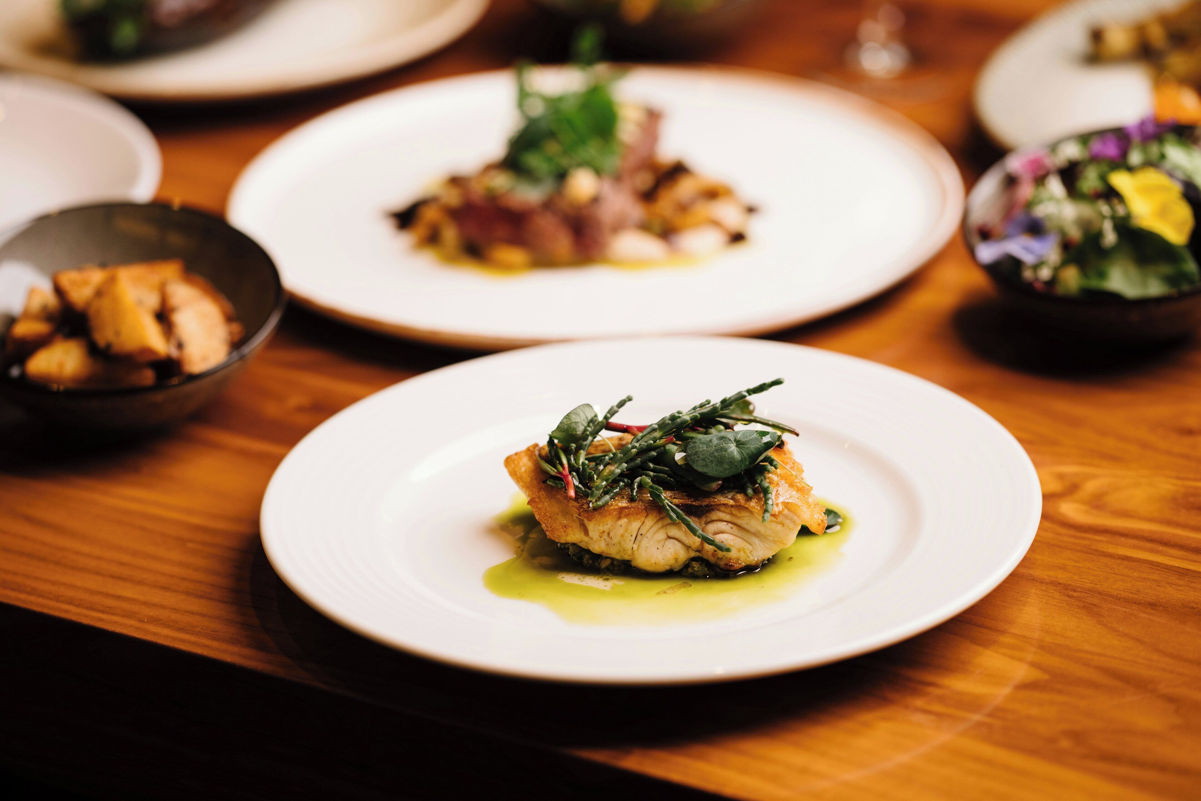 Une table à manger avec des assiettes remplies de nourriture, dont l’une contient un morceau de poisson garni de légumes verts.