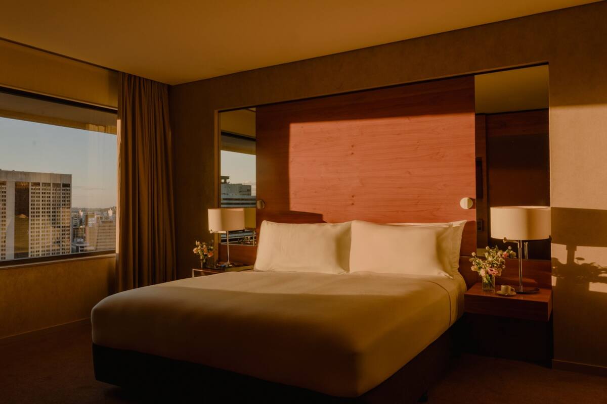 Habitación de hotel con una cama grande, una mesita de noche, una lámpara y vistas de la ciudad a través de un ventanal amplio.