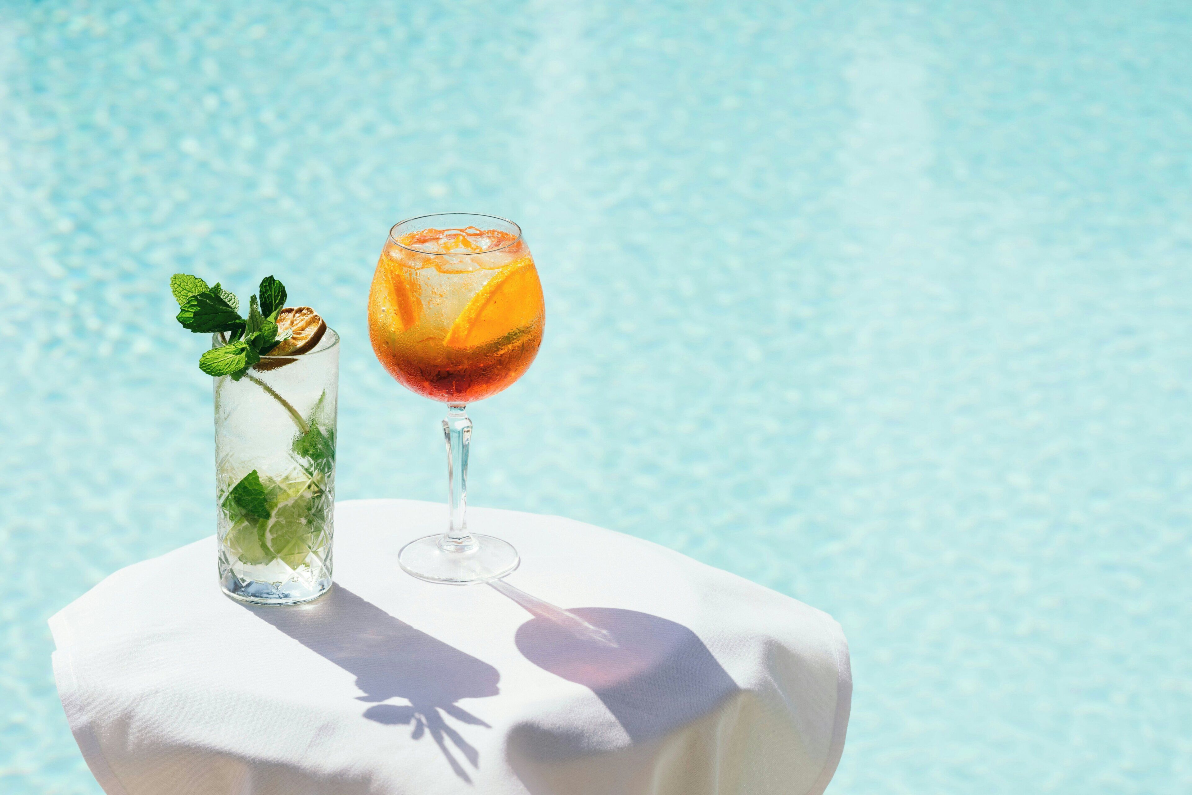 Two glasses of a beverage with mint leaves, one on a white surface and the other on a poolside table.