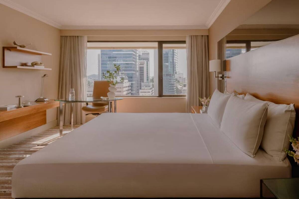 A hotel room with a large bed, a desk, a chair, a small table, and a view of the cityscape.