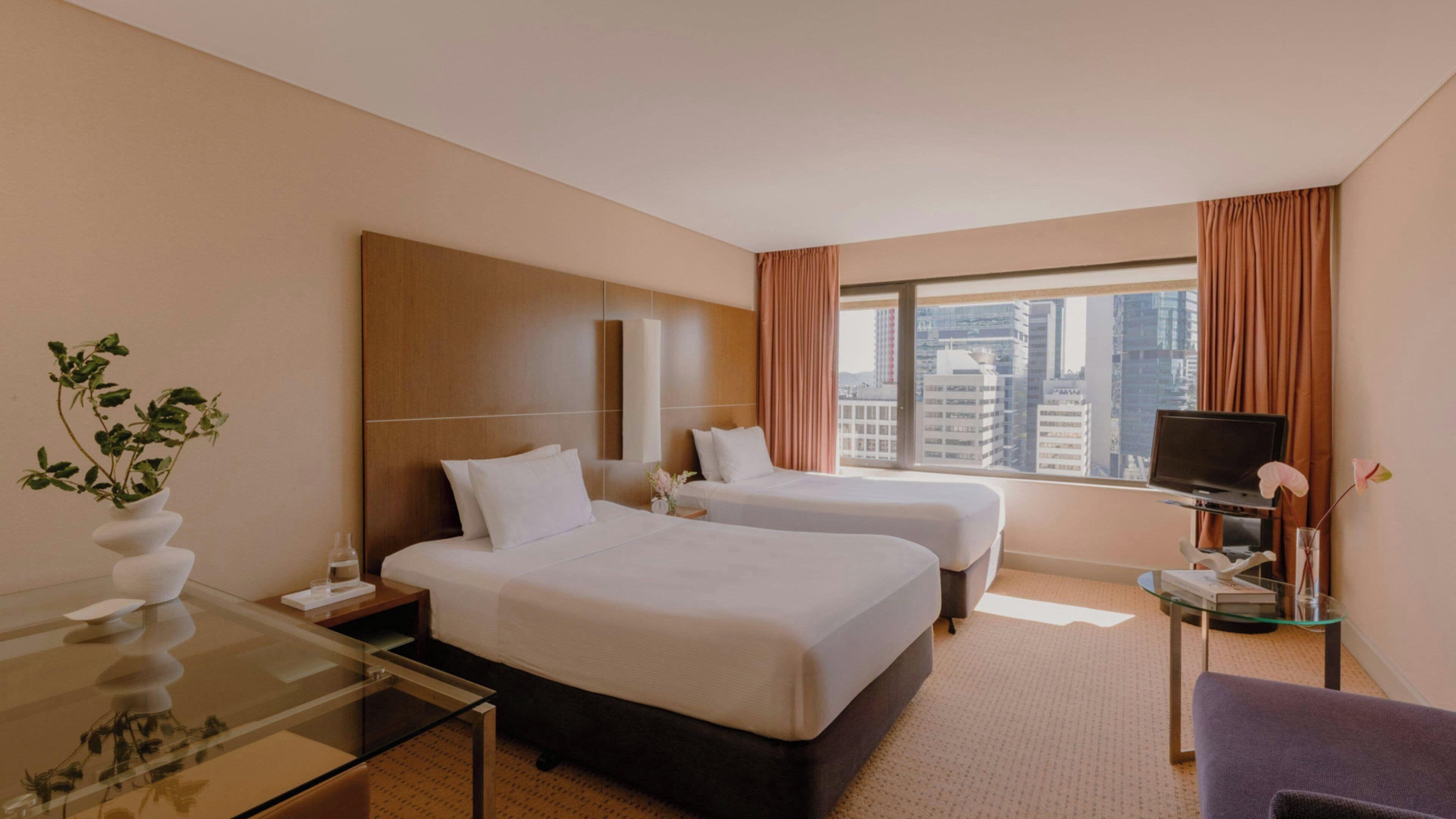 A hotel room with two beds, a TV, a glass table, and a view of the city.