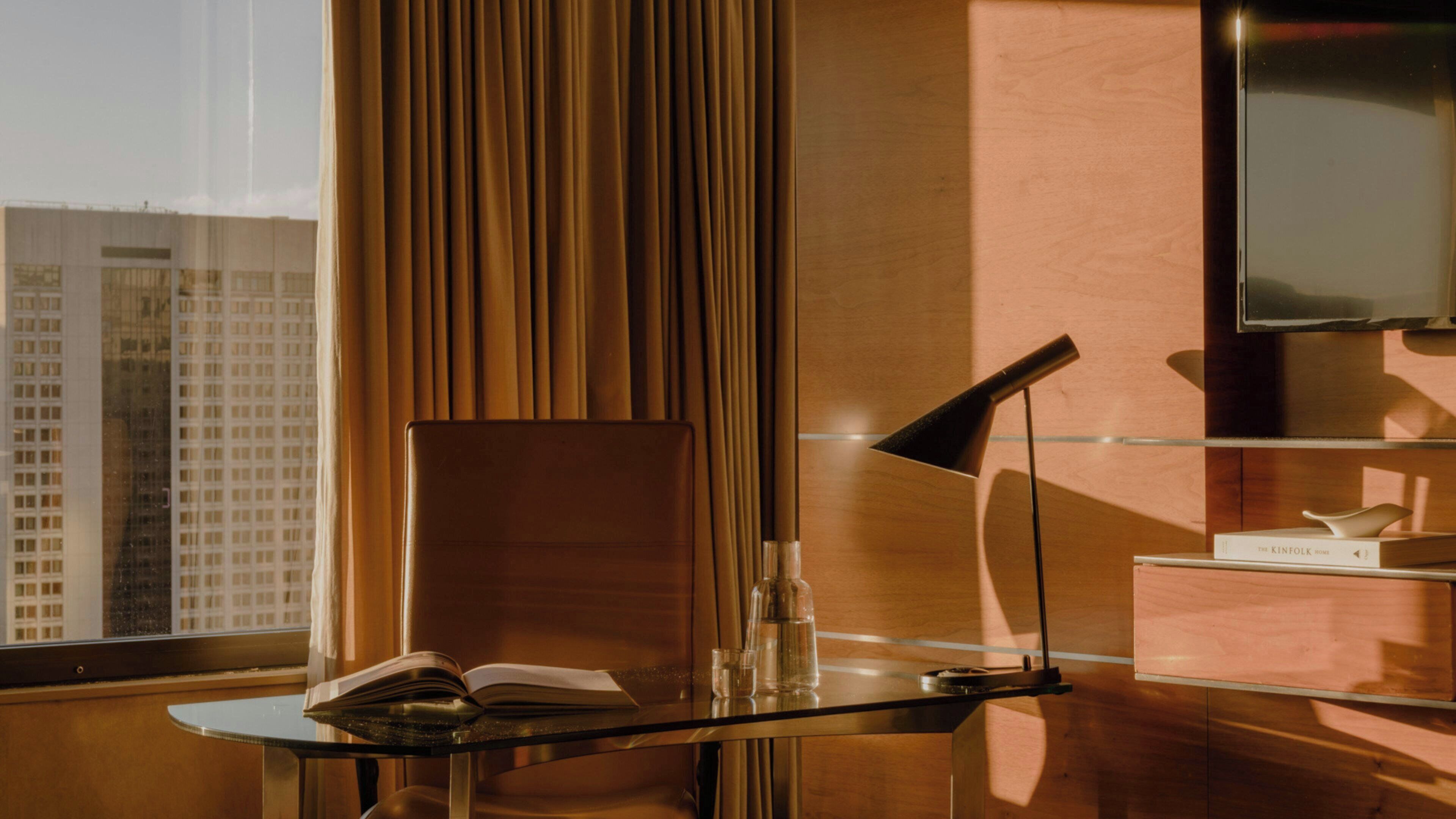 A modern hotel room with a glass table, a book, a lamp, and a view of a cityscape through a window with curtains.