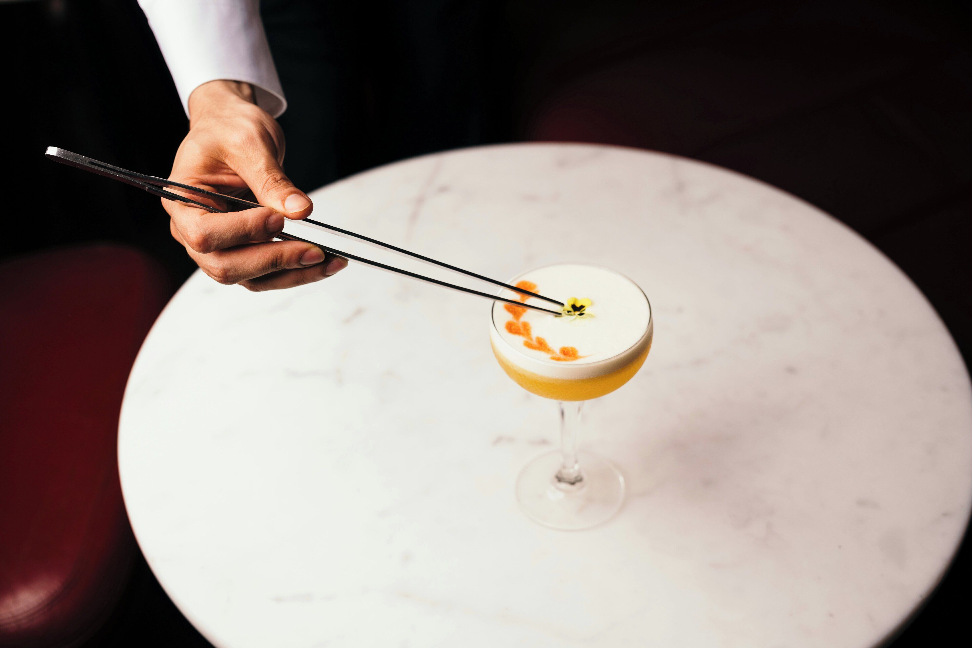A hand using chopsticks to pick up a cocktail with a layered drink and garnish on a white marble table.