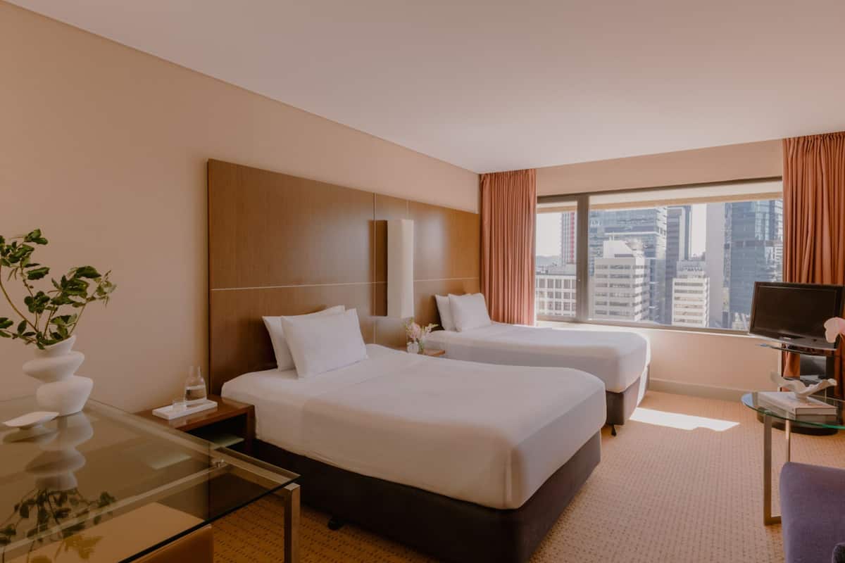 Habitación de hotel con dos camas, televisión, una mesa de vidrio y vistas a la ciudad.