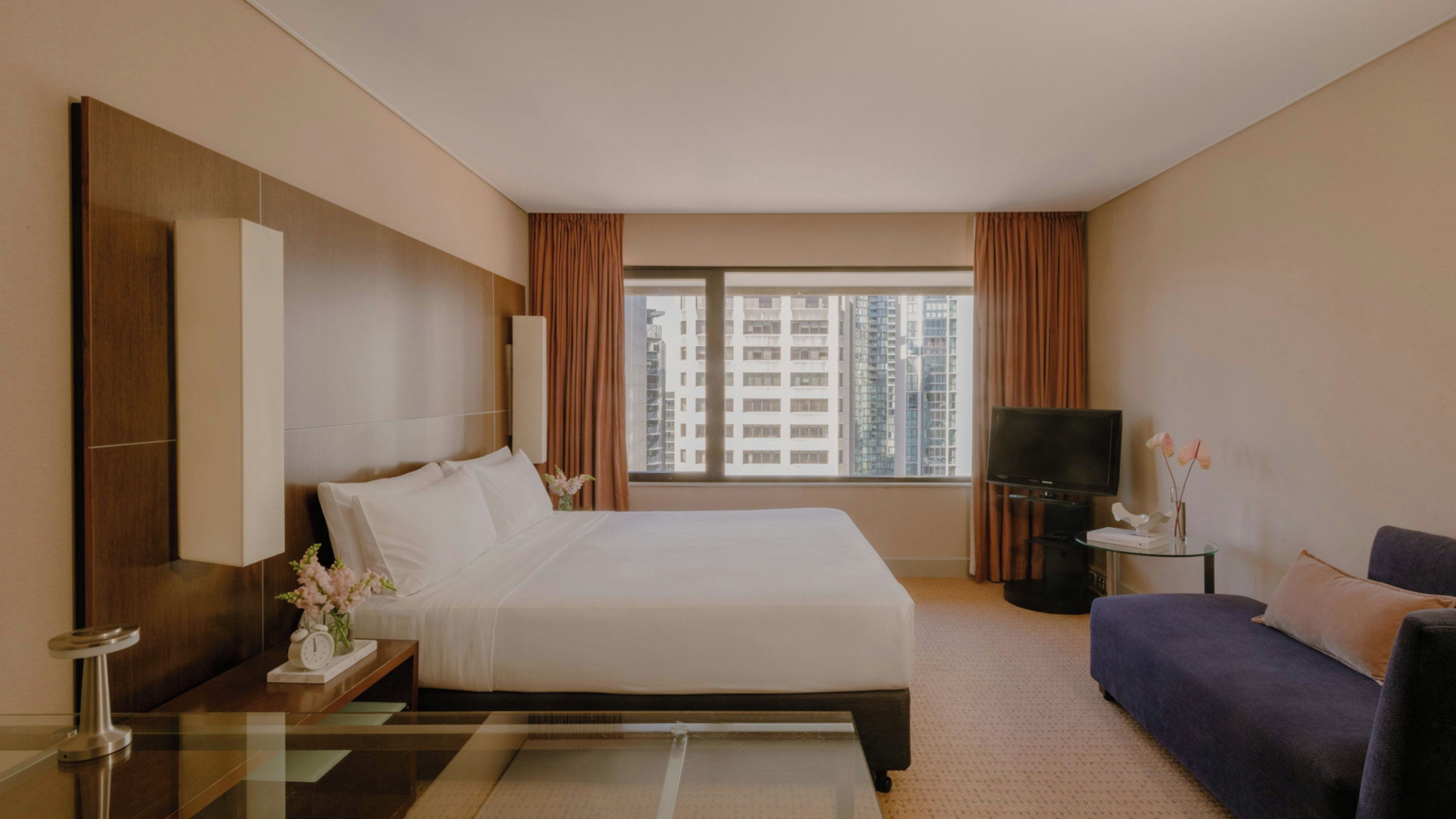 A hotel room with a large bed, a sofa, a TV, and a window with a city view.