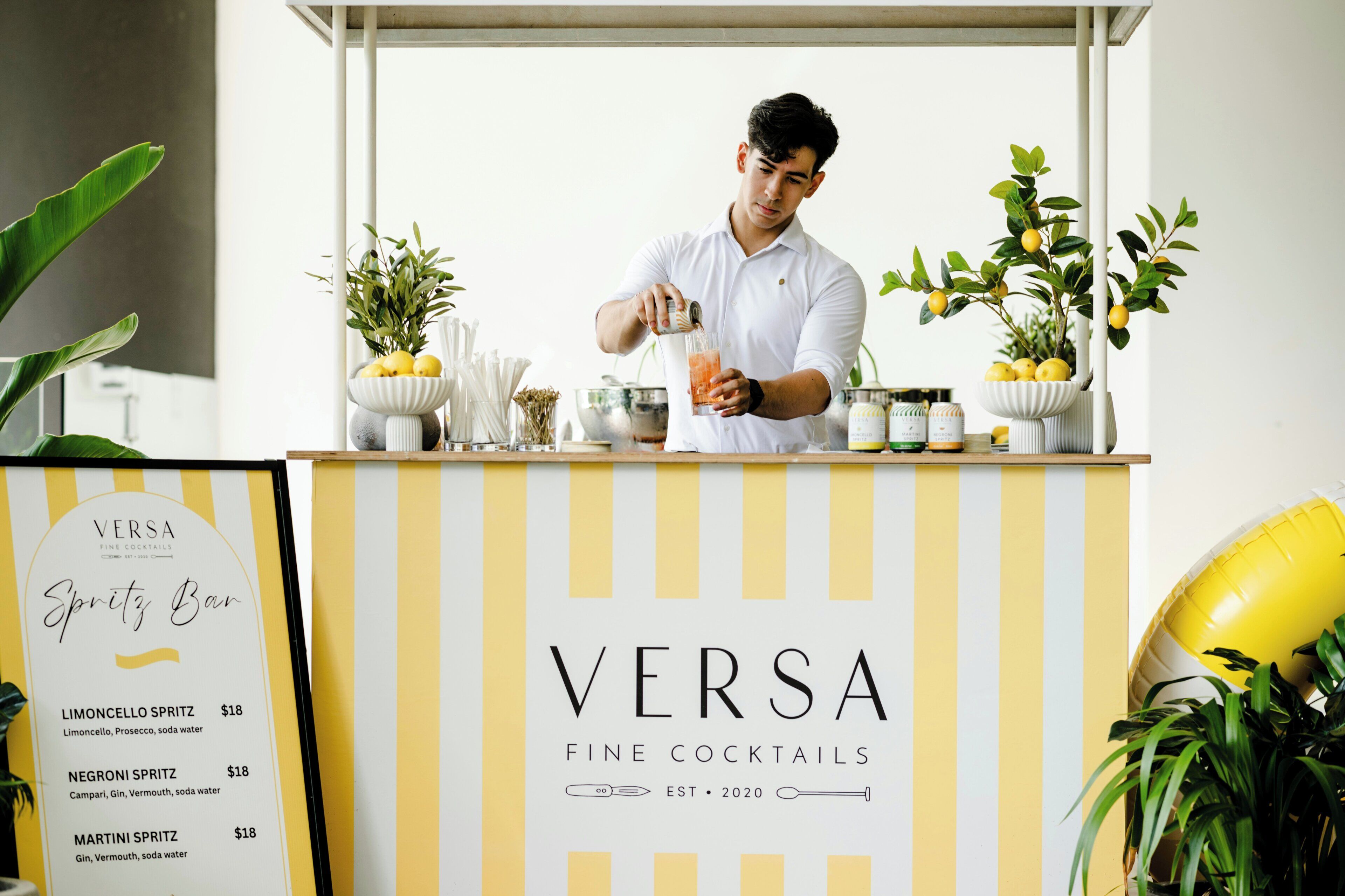 Ein Bartender hinter der Bar Versa Fine Cocktails, mit einer Getränkekarte, auf der Cocktails wie Limoncello Spritz und Martini Spritz angeboten werden.