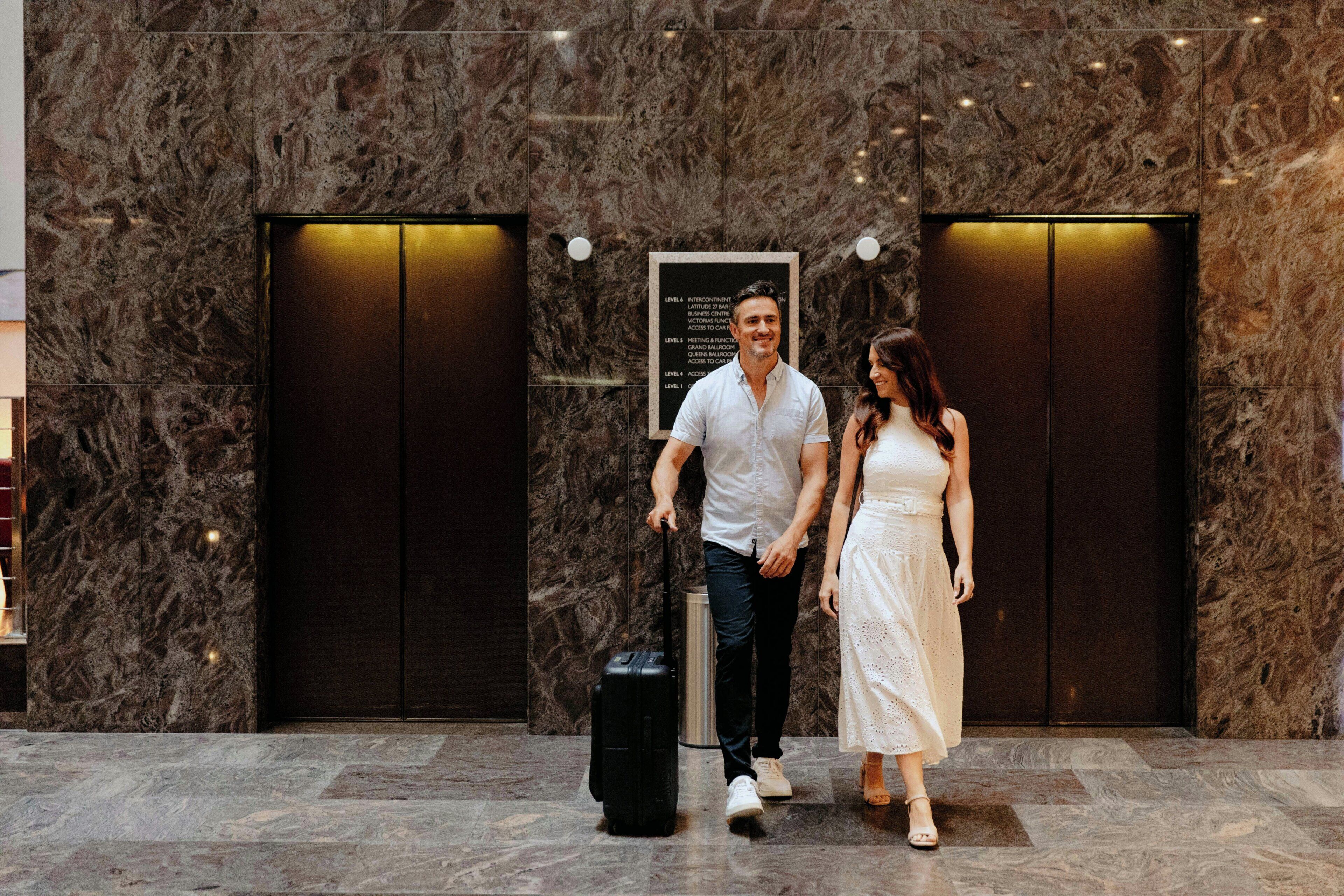 A man and a woman walking towards an elevator in a lobby with marble walls and two elevators.