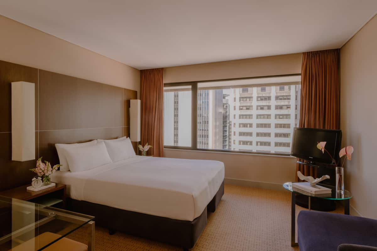Habitación de hotel con una cama grande, un escritorio con lámpara, televisión y vistas a edificios altos por la ventana.