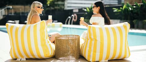 Dos personas sentadas sobre cojines a rayas amarillas y blancas junto a una piscina, con una cesta de mimbre entre ellas.