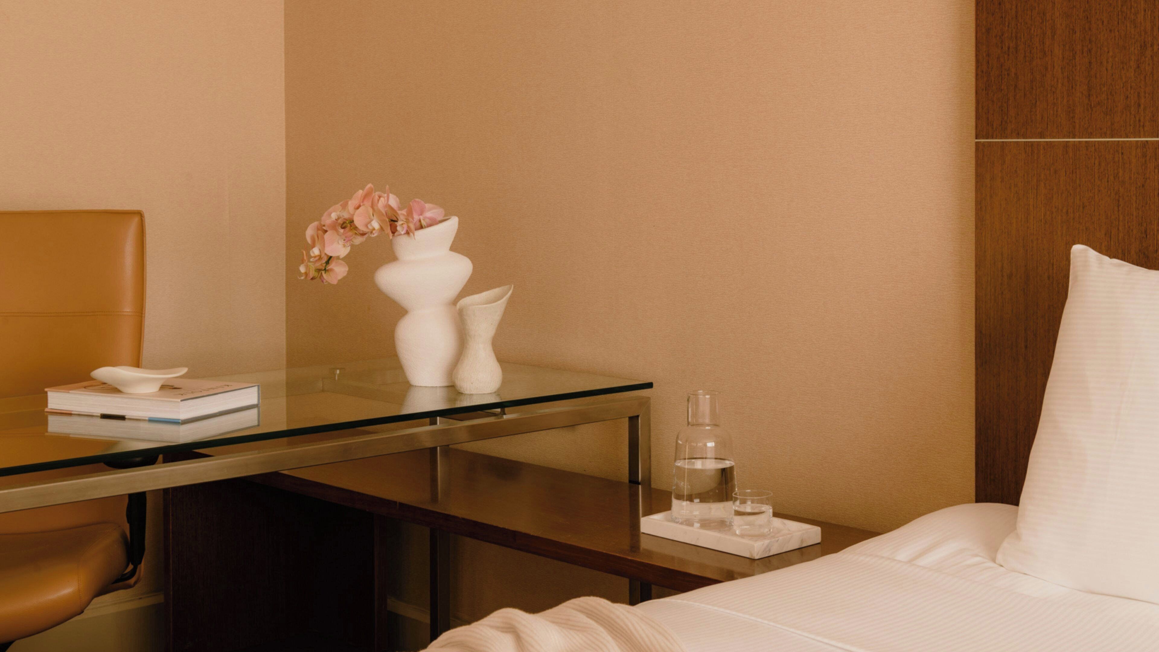 A hotel room with a bed, a desk with a vase of flowers, a book, and a water bottle.