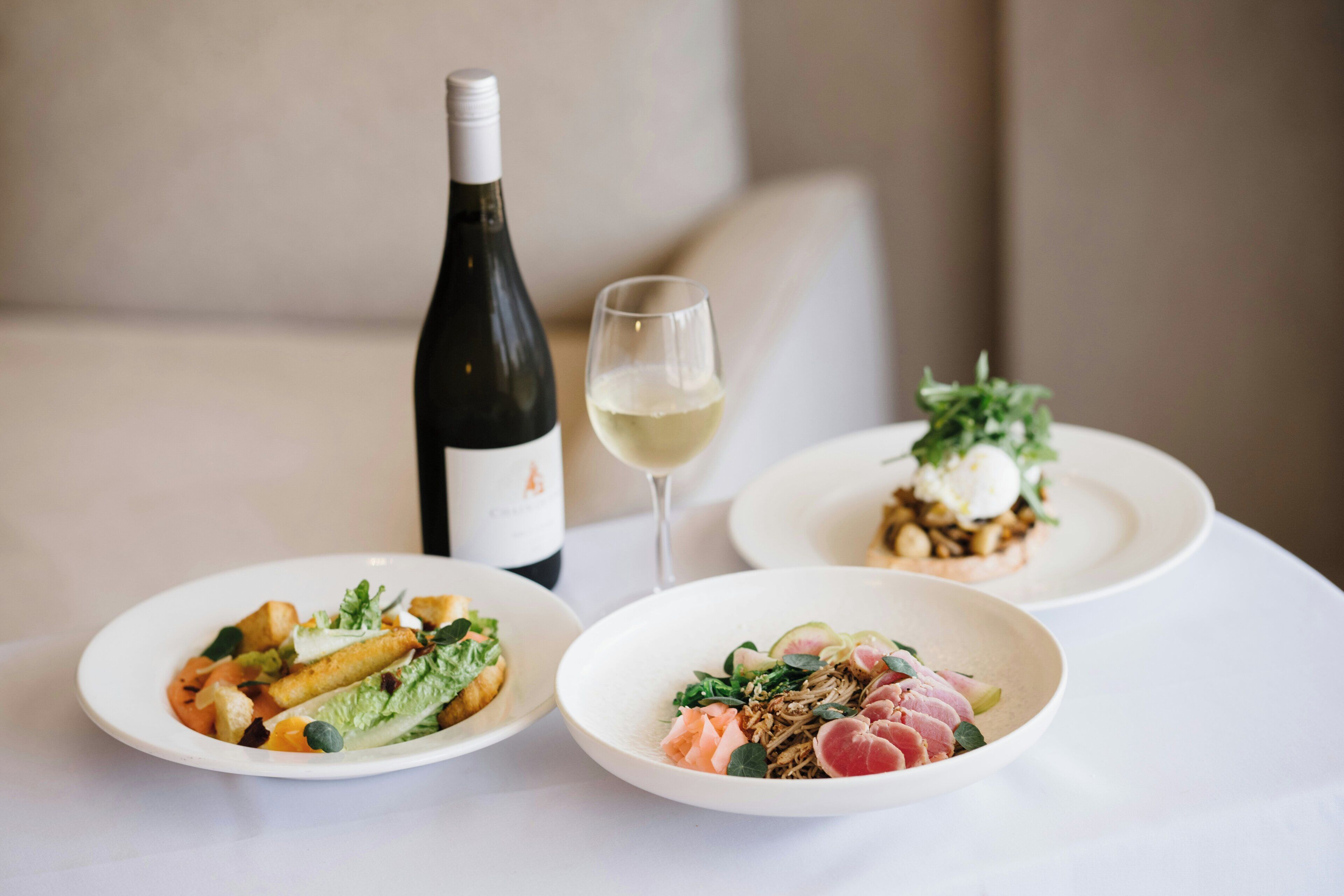 Three plates of food, a bottle of wine, and a glass of wine on a table.