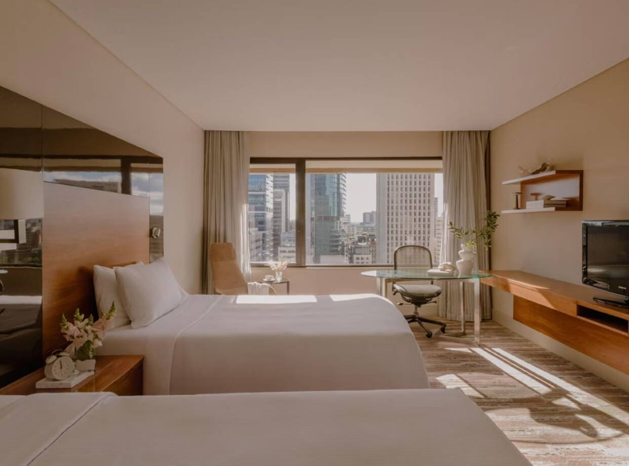 Habitación de hotel con dos camas, un escritorio, una silla, un televisor y un ventanal con vistas a la ciudad.