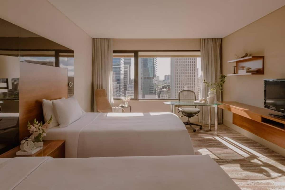 Habitación de hotel con dos camas, un escritorio, una silla, un televisor y un ventanal con vistas a la ciudad.