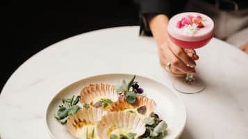 Una persona sosteniendo una copa con un cóctel rosado y un plato de comida con conchas de vieira y vegetales verdes.