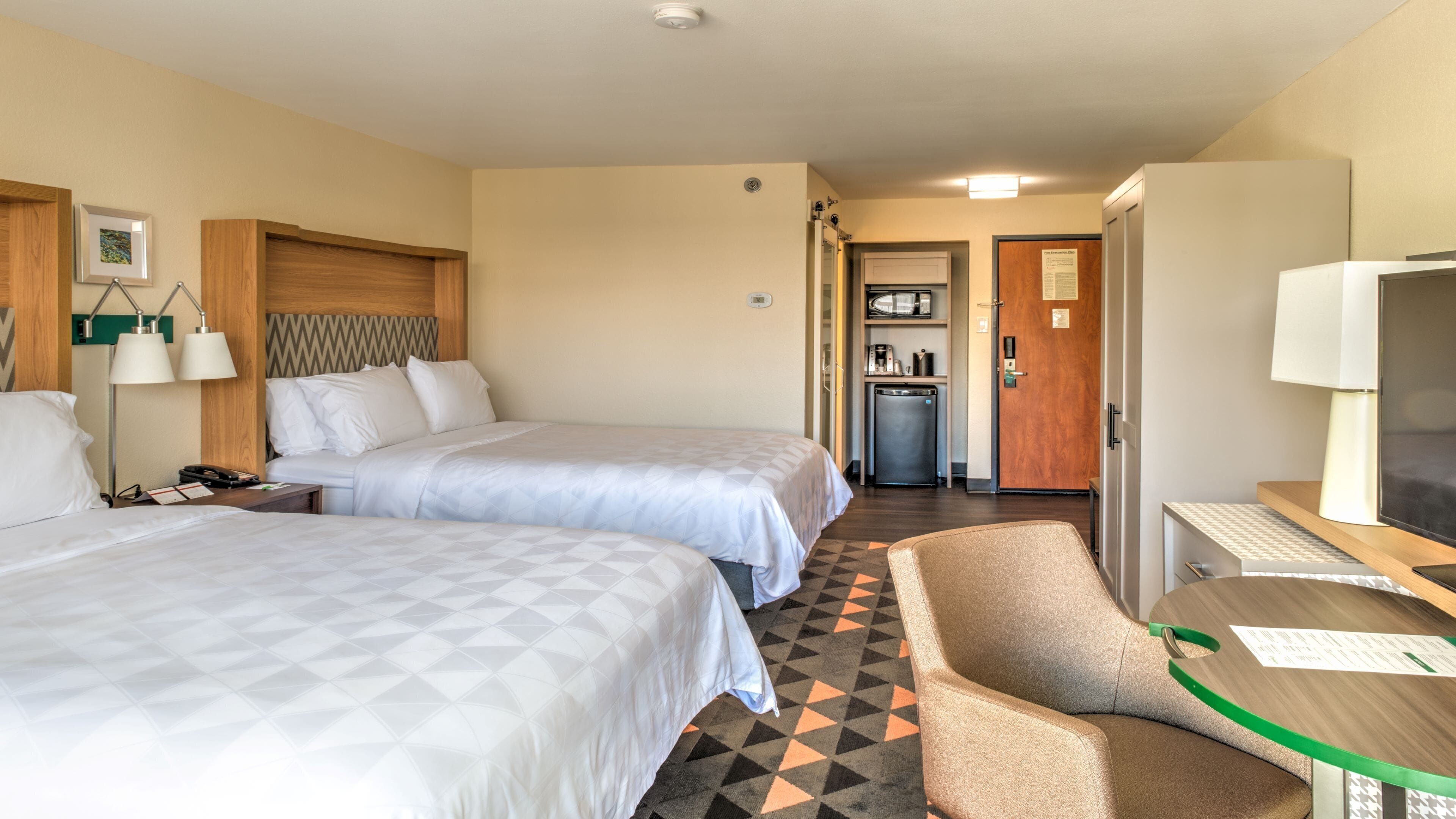 A hotel room with two beds, a desk, a chair, a television, and a kitchenette.