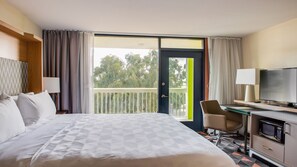 A hotel room with a bed, a desk with a television, a chair, and a balcony with a view of trees. - Holiday Inn Phoenix-Mesa/Chandler by IHG (Mesa)