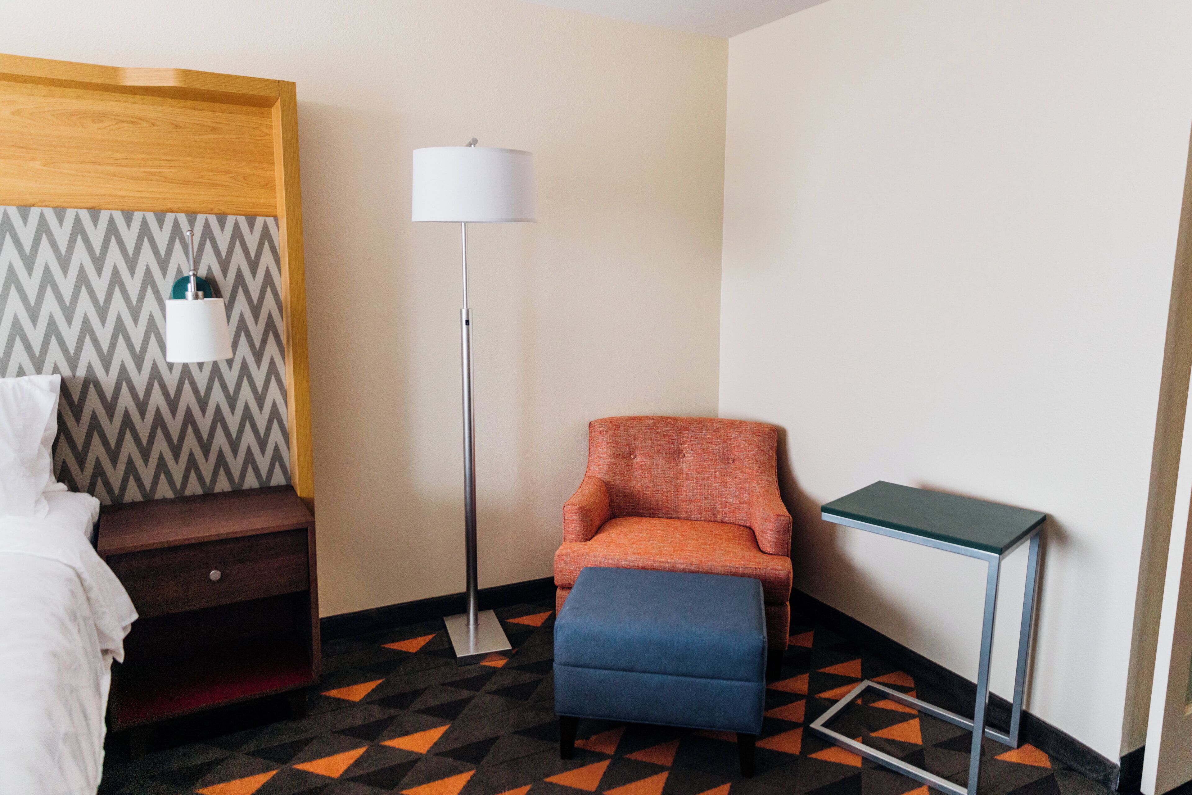 Una habitación de hotel con cama, mesita de noche, sillón con reposapiés, lámpara de pie y una pequeña mesa.