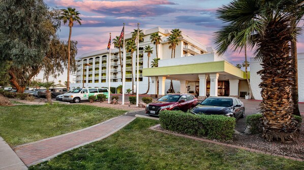 A hotel with a white facade, multiple balconies, and palm trees. There are several cars parked in front, and a pathway leading to the entrance. - Holiday Inn Phoenix-Mesa/Chandler by IHG (Mesa)