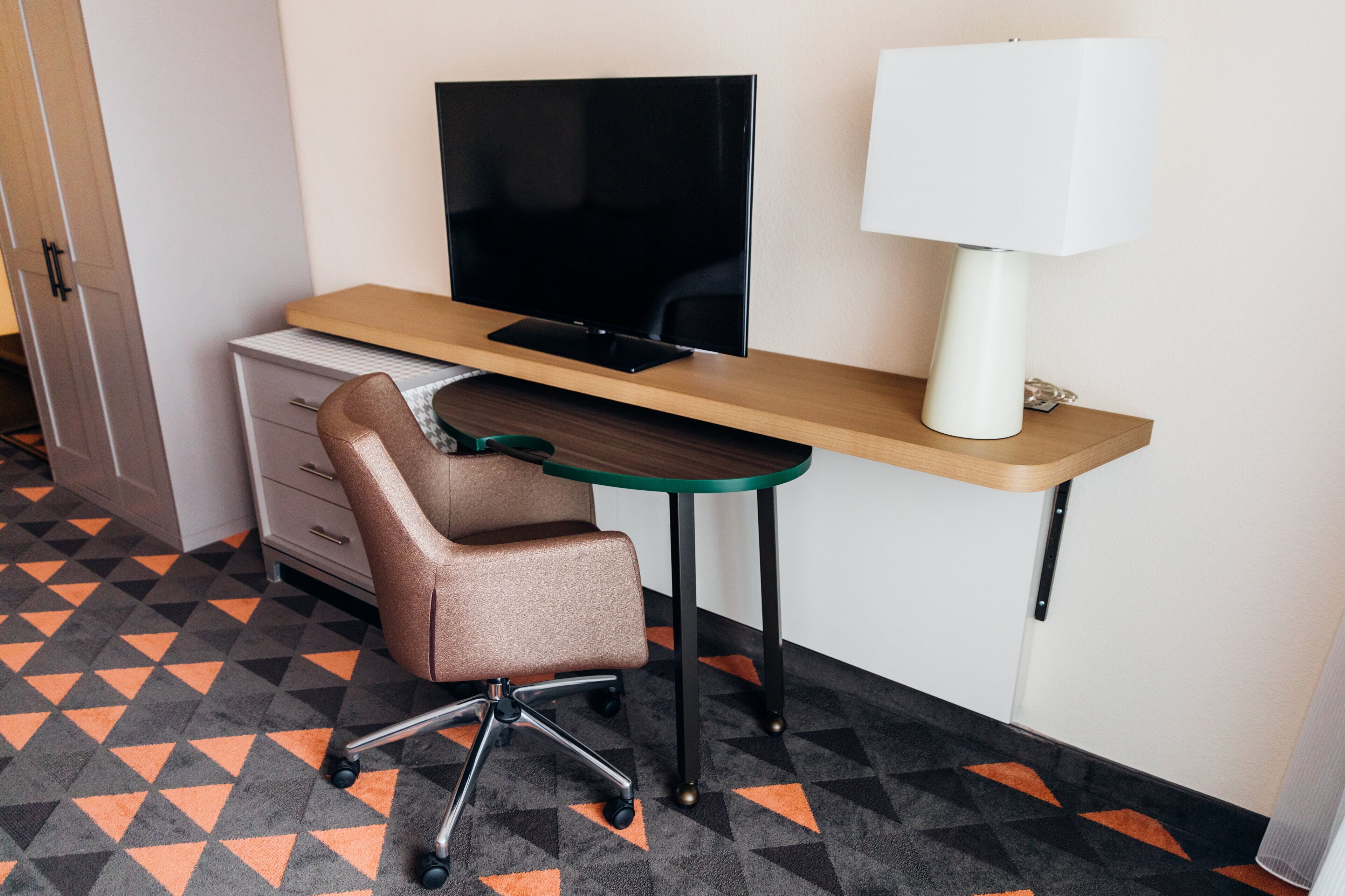 A hotel room with a desk, chair, TV, lamp, and patterned carpet.