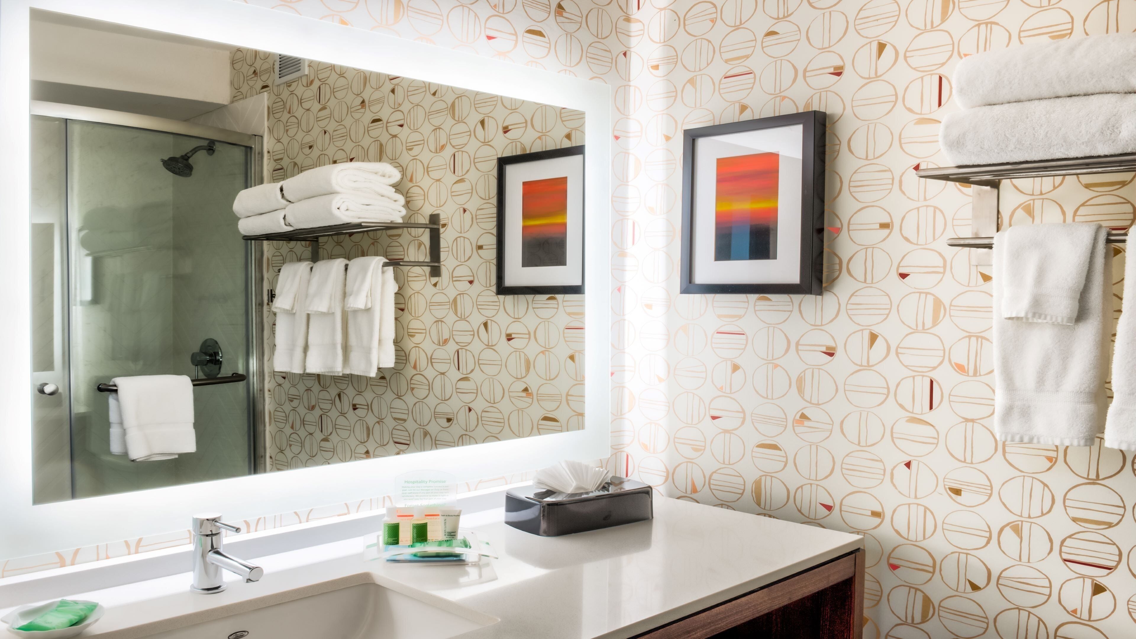 Un baño con un espejo grande, un lavabo, cabina de ducha de vidrio y toallas en un perchero. Hay cuadros en la pared.