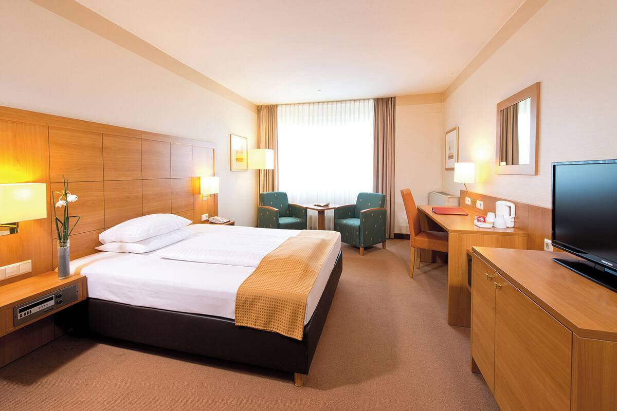 Ein Hotelzimmer mit Bett, Nachttischen, einem Schreibtisch mit Stuhl, einem Fernseher und einem Fenster mit Vorhängen.