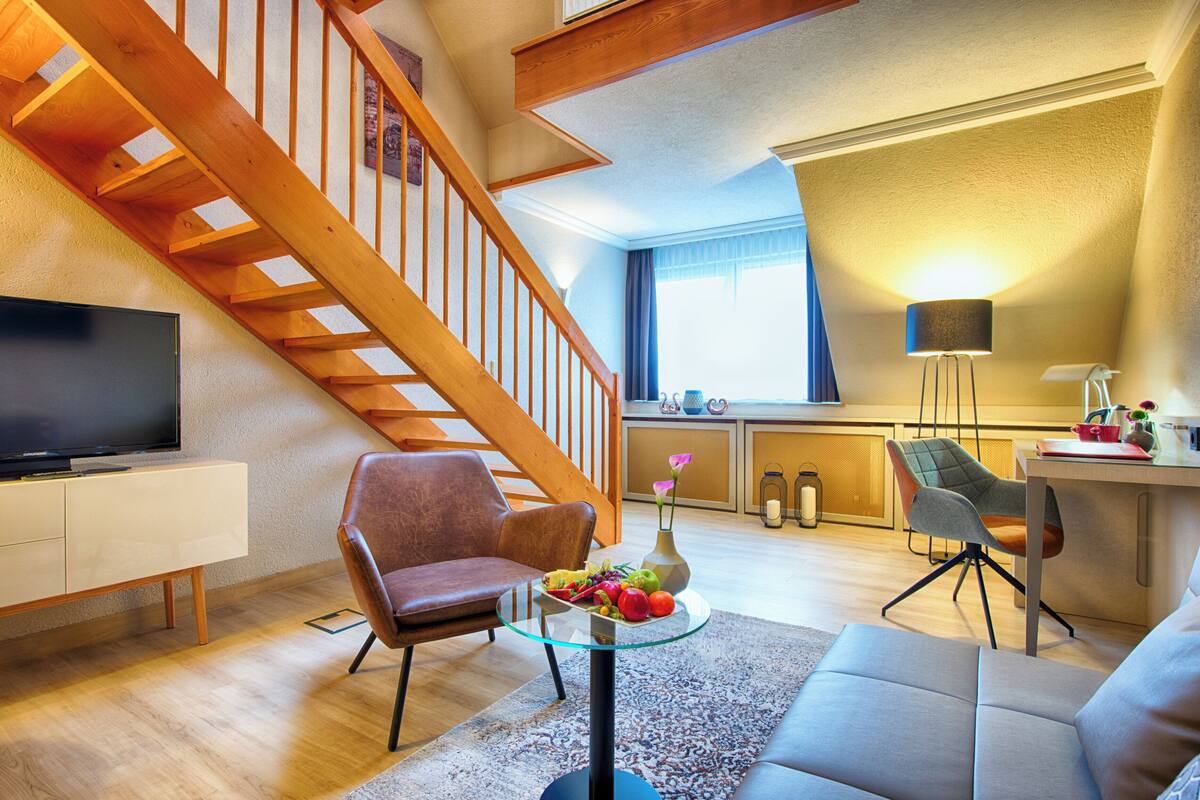 Ein modernes Wohnzimmer mit einer Holztreppe, einem Flachbildfernseher, einem Glastisch mit Obst und einer Couch.