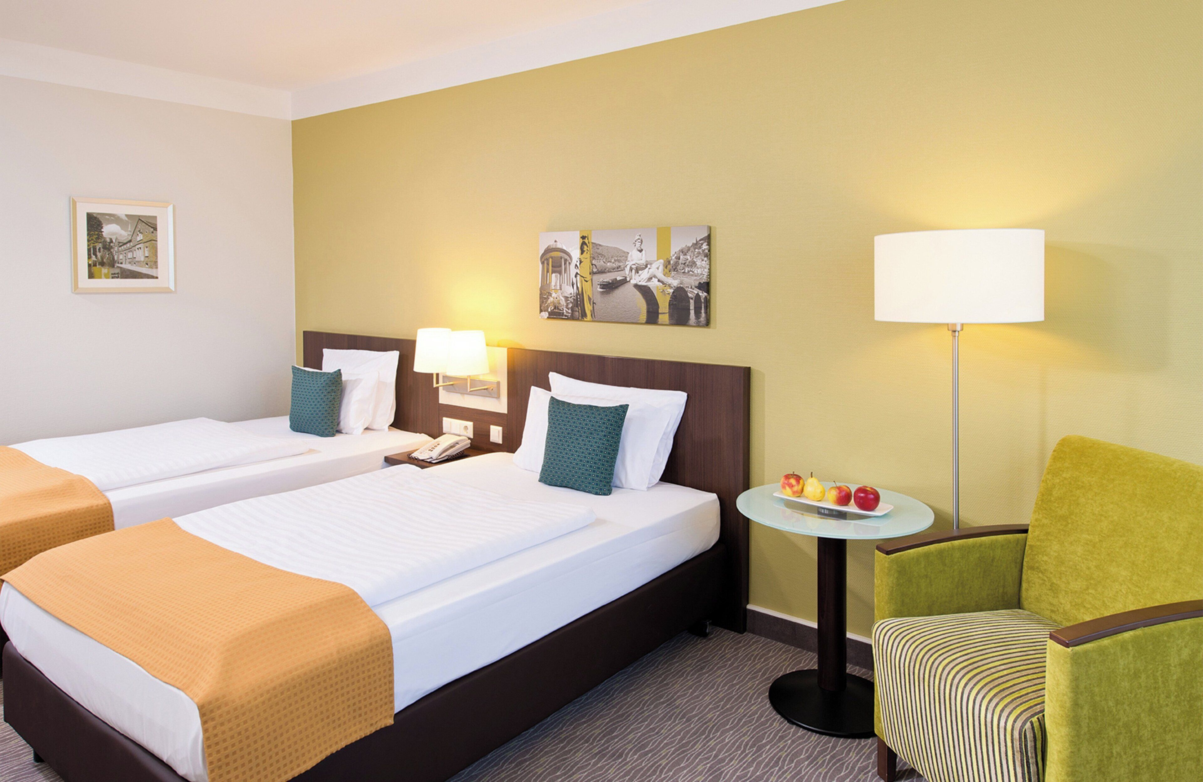 Quarto de hotel com duas camas, uma mesa redonda pequena com frutas, uma poltrona verde e um abajur.