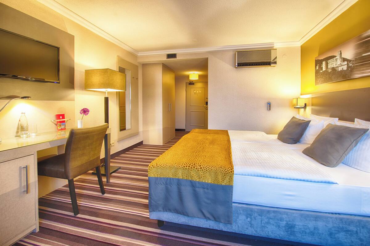 Quarto de hotel com cama, escrivaninha, cadeira, televisão e um quadro na parede.
