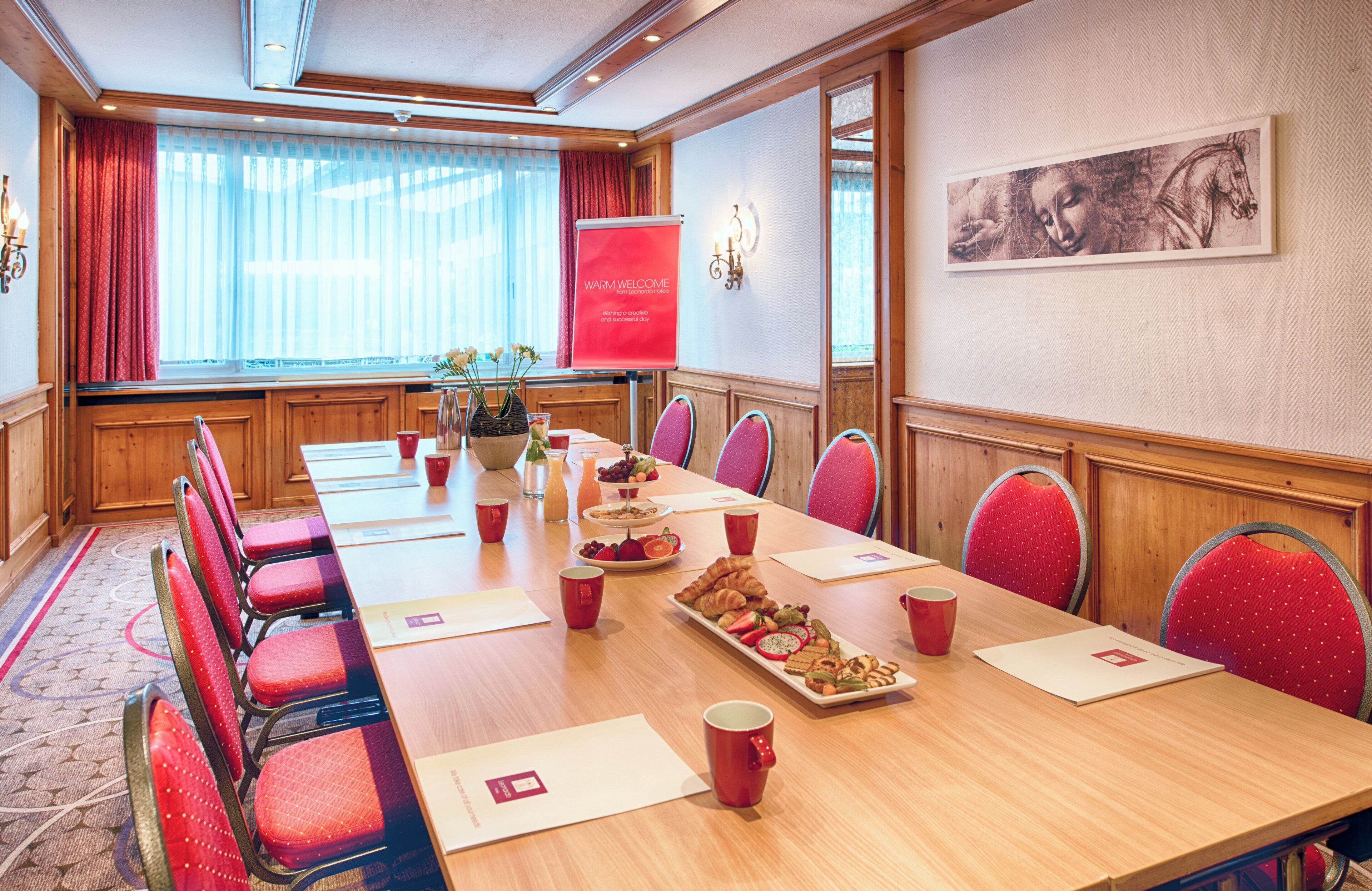 Ein Konferenzraum mit langem Tisch, roten Stühlen, einem roten Banner und einem gerahmten Kunstwerk an der Wand.