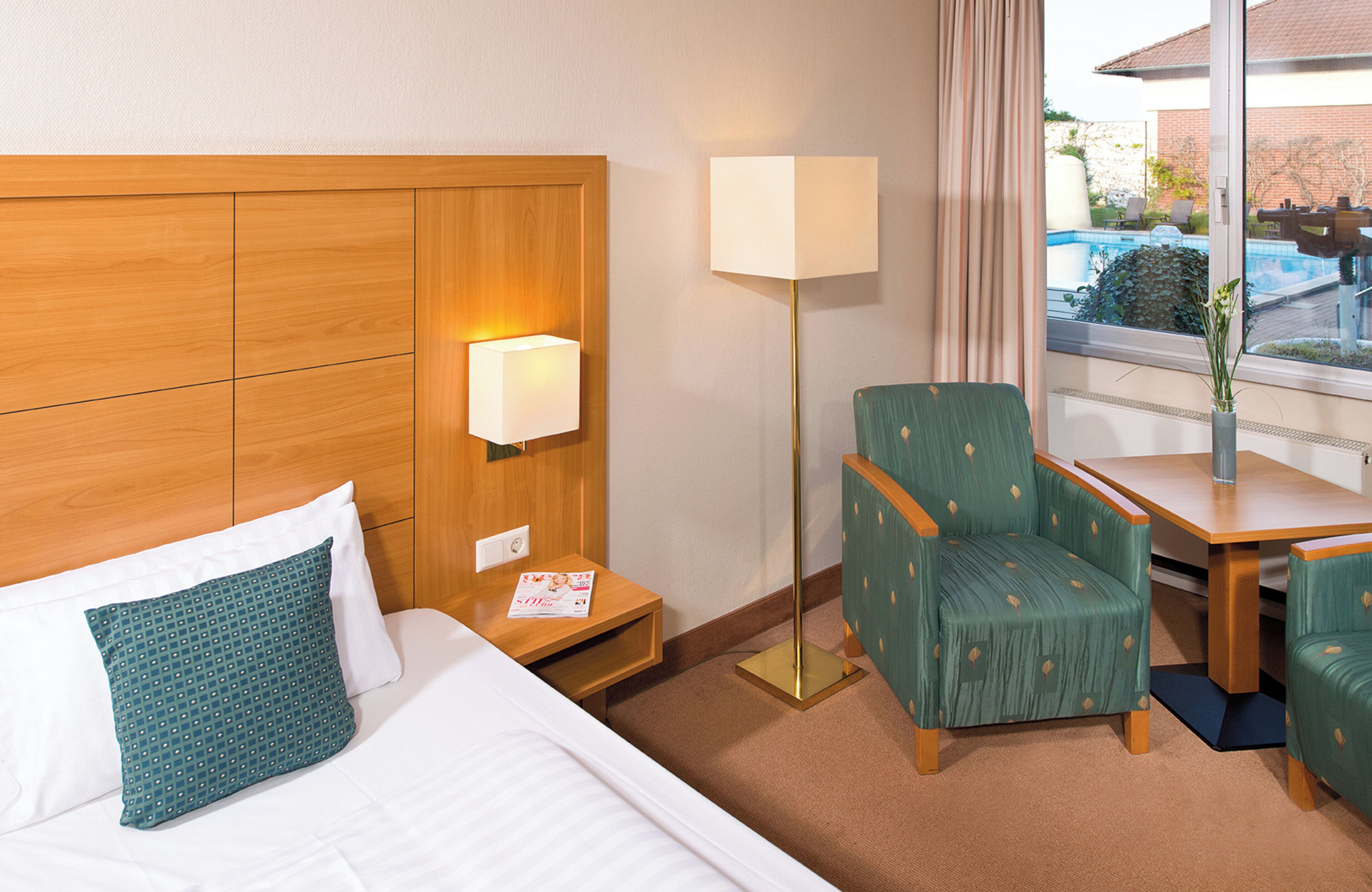 A hotel room with a wooden headboard, a bedside table with a lamp, a green armchair, a small table with a vase, and a view of a swimming pool through a window.