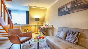 Una sala de estar moderna con un sofá de cuero marrón, una mesa de centro de vidrio con fruta, un sillón de cuero marrón, una escalera y una cocina al fondo.